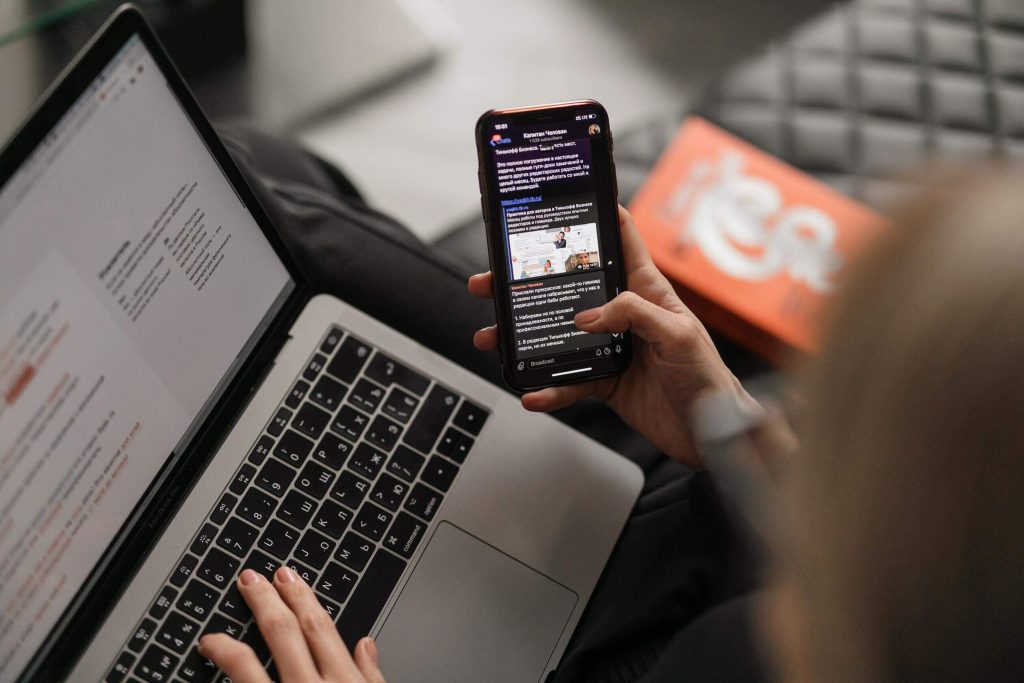 woman scrolling on her mobile phine while sitting comfortable on a couch in front of her laptop, showing different ways social engineering attacks can take place