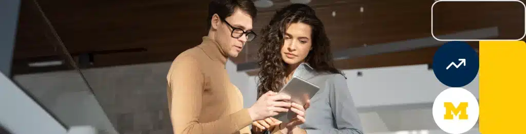 Two individuals using a tablet in a modern space with the University of Michigan logo